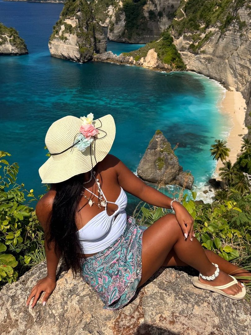 Vivian Dolor in a white top and sun hat sitting on a rock above Kelingking beach, Nusa Penida.